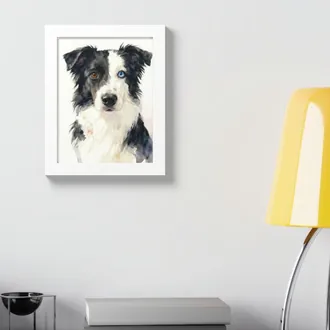 Framed watercolor dog portrait in a white frame on a light gray wall above a desk with a yellow lamp